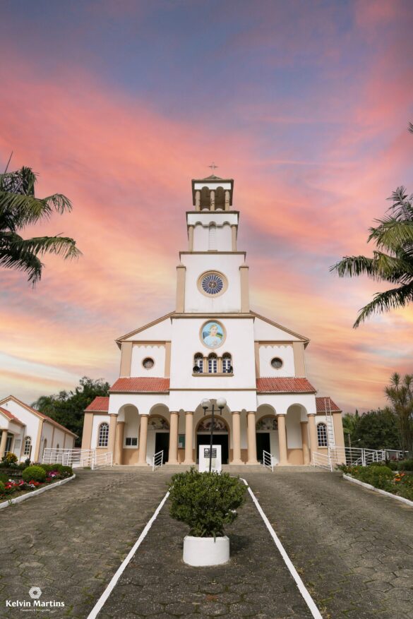 Fachada da igreja matriz de Imaruí com torre e relógio, palmeiras ao lado e céu com nuvens rosadas ao entardecer