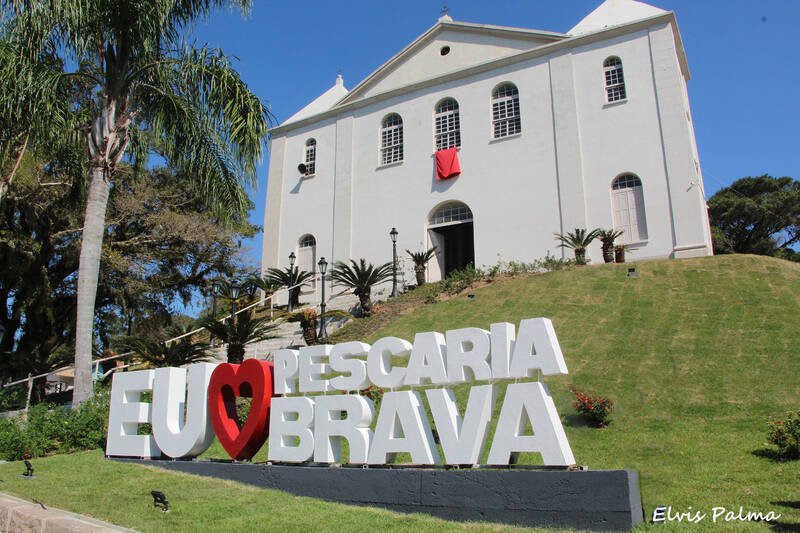 Igreja matriz branca do Senhor Bom Jesus do Socorro no centro de Pescaria Brava, Santa Catarina, com escadaria, faixa vermelha da festa do padroeiro e letreiro EU AMO PESCARIA BRAVA no jardim em frente