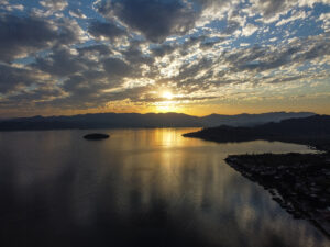 Vista aérea da Lagoa de Imaruí ao pôr do sol, com águas espelhadas, uma ilha e a Serra do Tabuleiro ao fundo, paisagem da rota turística SC-437 entre Pescaria Brava e Imaruí