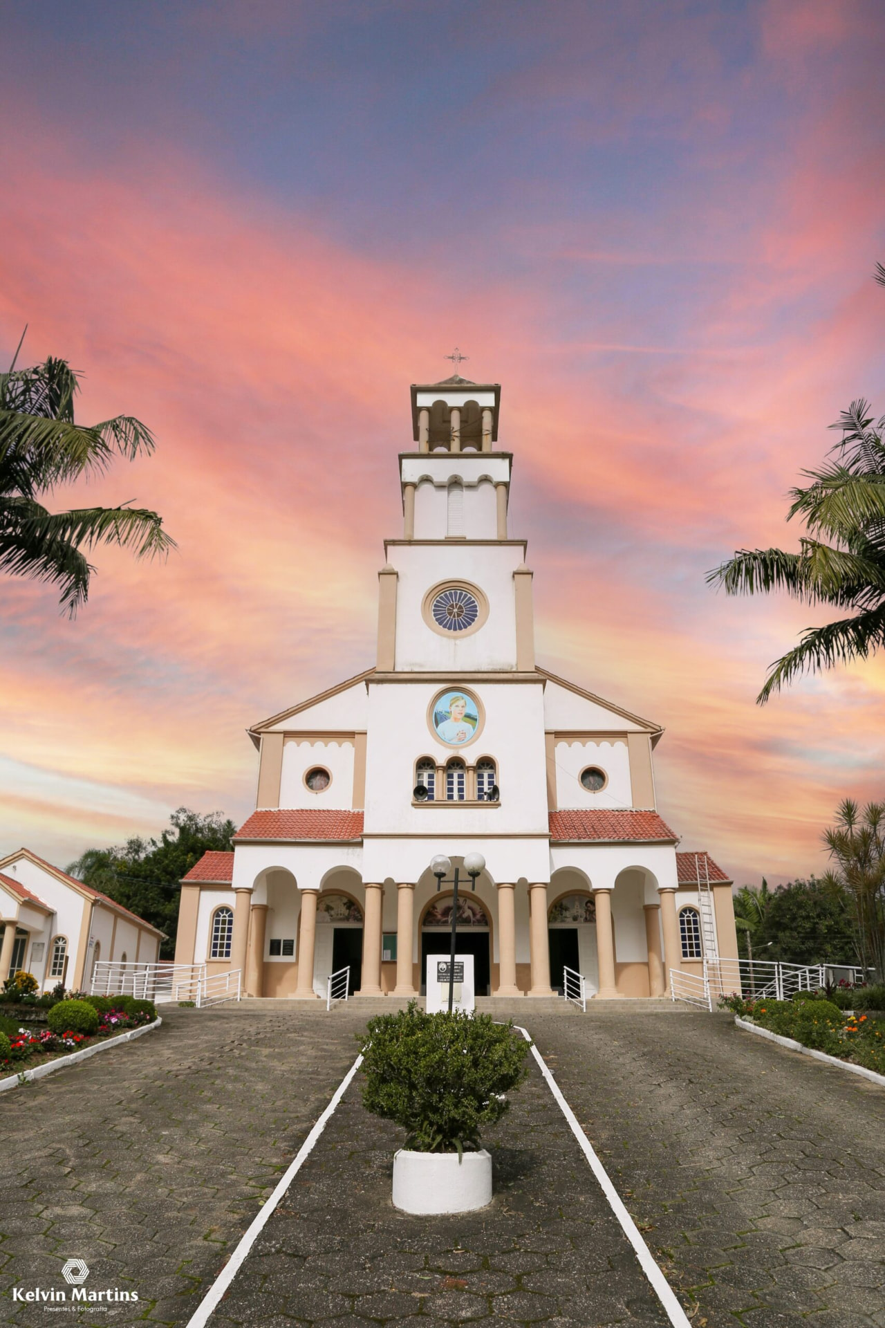 Torre branca do Santuário Diocesano da Bem-Aventurada Albertina Berkenbrock, em São Luís, Imaruí, com a imagem da beata na fachada, recortada contra um céu de pôr do sol rosa e laranja