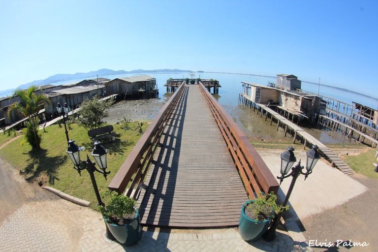Trapiche de madeira do porto de Pescaria Brava avançando sobre a Lagoa de Imaruí, com ranchos de pesca artesanal de madeira coloridos nas laterais e canoas atracadas, ponto inicial da rota turística SC-437 em Santa Catarina